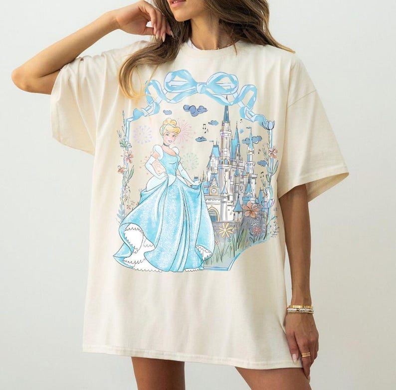 May include: Cream-colored t-shirt featuring a Cinderella graphic. The design includes Cinderella in a blue gown, a castle, and a blue bow. The shirt has short sleeves and a relaxed fit.