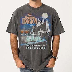 May include: Dark gray t-shirt featuring a graphic from "The Haunted Mansion" ride at Disneyland. The design includes the ride's logo, a spooky mansion, and ghostly figures. The text "They're Dying To Meet You!" and "Fantasyland" are also visible.
