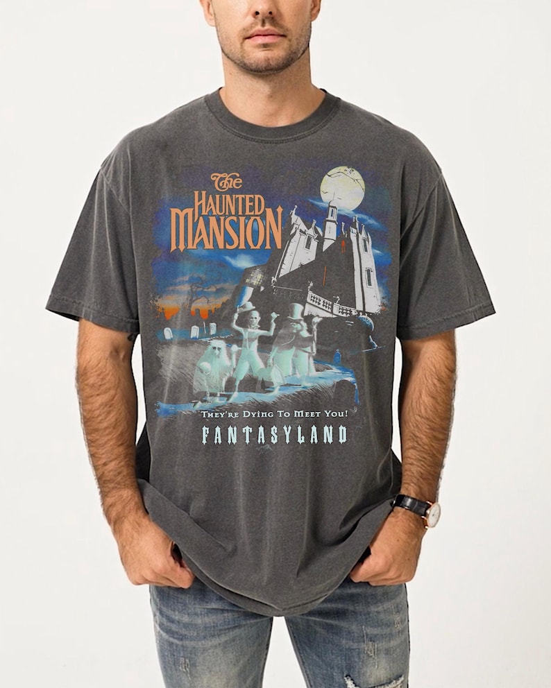 May include: Dark gray t-shirt featuring a graphic from "The Haunted Mansion" ride at Disneyland. The design includes the ride's logo, a spooky mansion, and ghostly figures. The text "They're Dying To Meet You!" and "Fantasyland" are also visible.