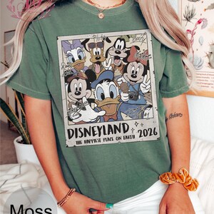 K&ouml;nnte beinhalten: Moosgr&uuml;nes T-Shirt mit einem Disneyland-Motiv mit Micky Maus, Donald Duck und anderen Disney-Figuren. Der Aufdruck enth&auml;lt den Text "Disneyland 2026" und "The Happiest Place on Earth."