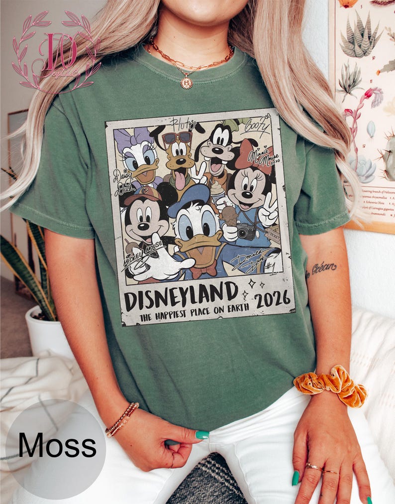 K&ouml;nnte beinhalten: Moosgr&uuml;nes T-Shirt mit einem Disneyland-Motiv mit Micky Maus, Donald Duck und anderen Disney-Figuren. Der Aufdruck enth&auml;lt den Text "Disneyland 2026" und "The Happiest Place on Earth."