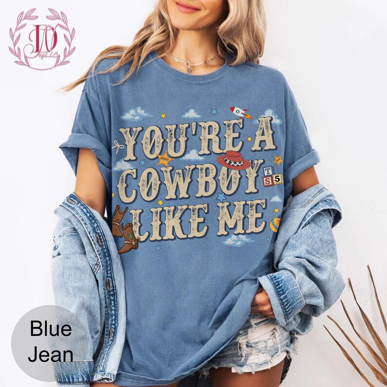 May include: A blue short-sleeve t-shirt featuring the text "YOU'RE A COWBOY LIKE ME" in a large, decorative font. The design includes small illustrations of a rocket, a cowboy hat, a pair of boots, stars, clouds, and blocks with the letters T, S, and 5.