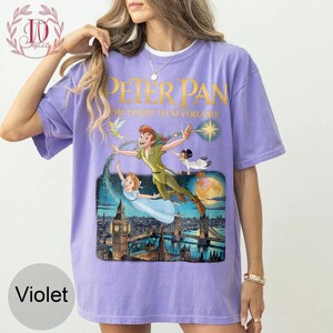 May include: A violet t-shirt with a graphic of Peter Pan, Wendy, Tinkerbell, and other characters flying over London. The text "PETER PAN" and "THE FLIGHT TO NEVERLAND!" are printed in gold. The shirt has a crew neck and short sleeves.