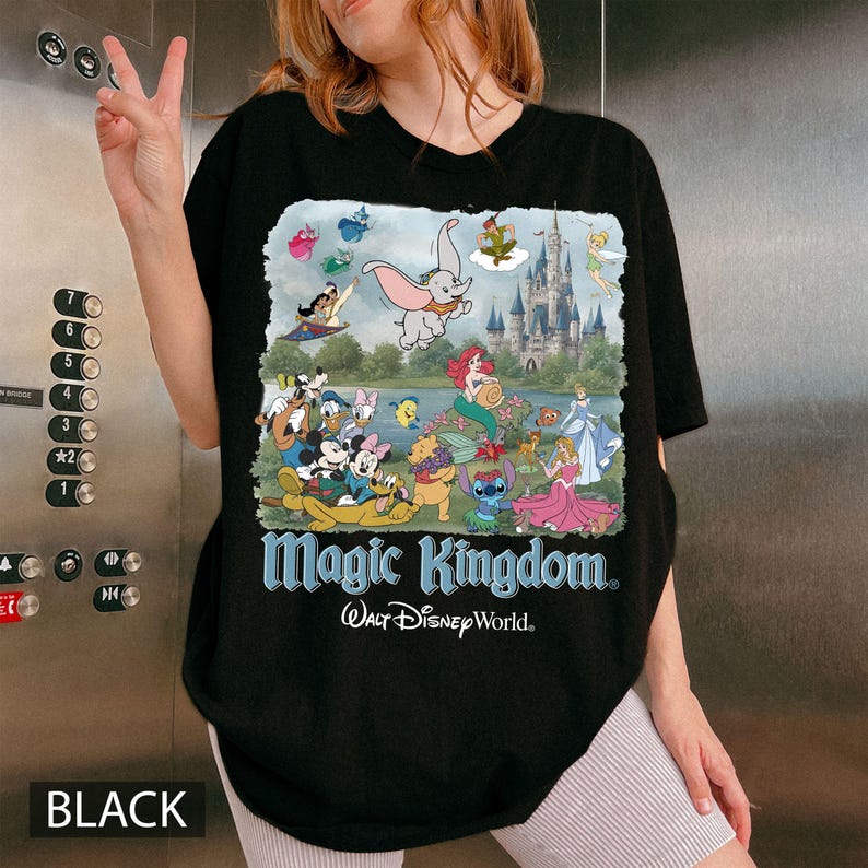 May include: A black t-shirt featuring a colorful graphic print of various animated characters, a castle, and the text Magic Kingdom and Walt Disney World. The design includes Dumbo, Ariel, Winnie the Pooh, Stitch, and other figures arranged in a scene.