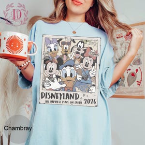 K&ouml;nnte beinhalten: Hellblaues T-Shirt mit einem Disneyland-Motiv im Vintage-Stil mit Micky Maus, Donald Duck und anderen Disney-Figuren. Der Aufdruck enth&auml;lt den Text "Disneyland The Happiest Place on Earth 2026".