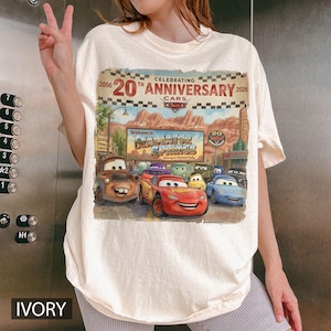 Puede incluir: Una camiseta de color marfil con un estampado gr&aacute;fico que celebra el 20 aniversario de la pel&iacute;cula Cars. El dise&ntilde;o muestra personajes como Rayo McQueen y Mate frente a un letrero de Radiator Springs con el texto 2006 20th Anniversary 2026.