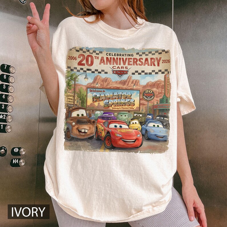 Puede incluir: Una camiseta de color marfil con un estampado gr&aacute;fico que celebra el 20 aniversario de la pel&iacute;cula Cars. El dise&ntilde;o muestra personajes como Rayo McQueen y Mate frente a un letrero de Radiator Springs con el texto 2006 20th Anniversary 2026.