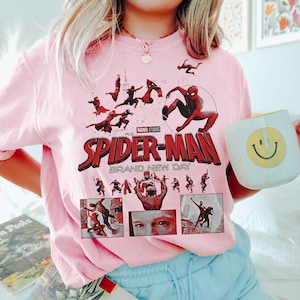 May include: A light pink sweatshirt featuring a graphic of Spider-Man with the text "SPIDER-MAN BRAND NEW DAY" in red and white. The design includes multiple images of Spider-Man in action. A white mug with a yellow smiley face is held in the image.