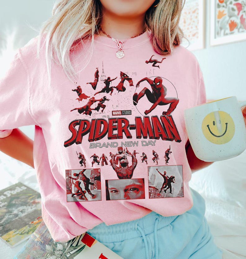 May include: A light pink sweatshirt featuring a graphic of Spider-Man with the text "SPIDER-MAN BRAND NEW DAY" in red and white. The design includes multiple images of Spider-Man in action. A white mug with a yellow smiley face is held in the image.