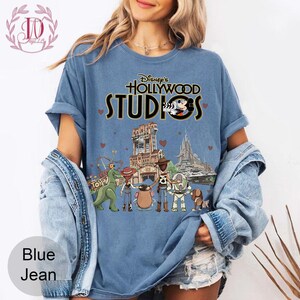 Pode incluir: Camiseta azul jeans com a frase "Disney's Hollywood Studios" e Mickey Mouse. O gr&aacute;fico inclui a Torre do Terror e outras atra&ccedil;&otilde;es do parque. A camiseta tem mangas curtas e um corte folgado.
