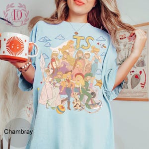 May include: A light blue t-shirt featuring a graphic print of multiple animated characters in various outfits, including a cowgirl, a space ranger, and a shepherdess. The design includes the letters TS, clouds, a rocket, and a ball. The shirt has short sleeves and a crew neckline.