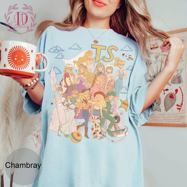 May include: A light blue t-shirt featuring a graphic print of multiple animated characters in various outfits, including a cowgirl, a space ranger, and a shepherdess. The design includes the letters TS, clouds, a rocket, and a ball. The shirt has short sleeves and a crew neckline.