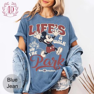 K&ouml;nnte beinhalten: Blaues Jeans-T-Shirt mit einem Mickey Mouse-Motiv und dem Text "LIFE'S A WALK IN THE PARK" und "Walt Disney World". Das Design beinhaltet ein Schloss und Feuerwerk. Das T-Shirt hat kurze &Auml;rmel.