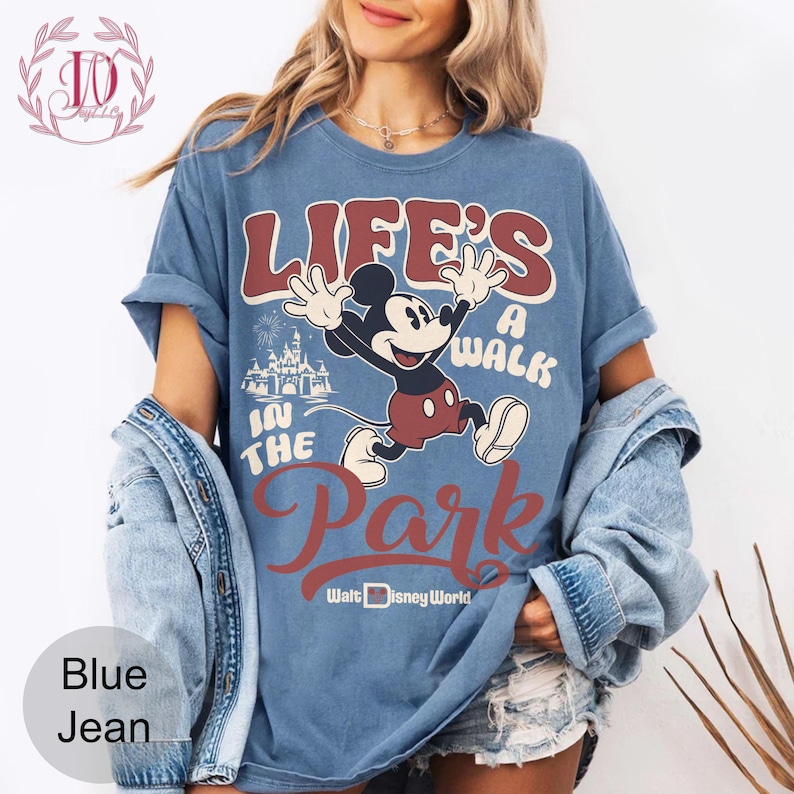 K&ouml;nnte beinhalten: Blaues Jeans-T-Shirt mit einem Mickey Mouse-Motiv und dem Text "LIFE'S A WALK IN THE PARK" und "Walt Disney World". Das Design beinhaltet ein Schloss und Feuerwerk. Das T-Shirt hat kurze &Auml;rmel.