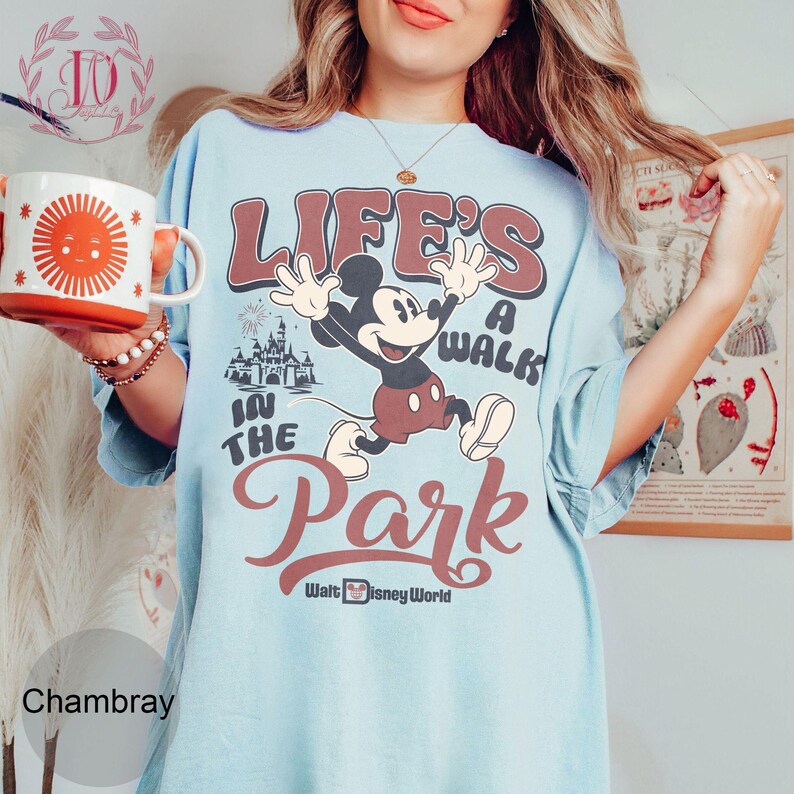 K&ouml;nnte beinhalten: Hellblaues T-Shirt mit einer Grafik von Mickey Mouse, der springt, und dem Text "Life's a Walk in the Park" und "Walt Disney World". Eine wei&szlig;-orange Tasse mit einem Sonnendesign wird in der linken Hand gehalten.