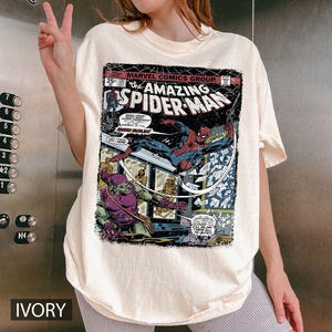 May include: Ivory-colored t-shirt featuring a vintage comic book cover of "The Amazing Spider-Man." The graphic showcases Spider-Man in action, battling a villain. The shirt includes the Marvel Comics Group logo and the number 137.