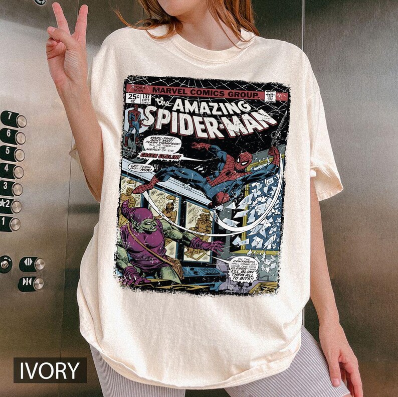 May include: Ivory-colored t-shirt featuring a vintage comic book cover of "The Amazing Spider-Man." The graphic showcases Spider-Man in action, battling a villain. The shirt includes the Marvel Comics Group logo and the number 137.