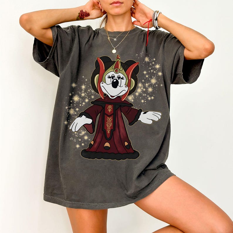 Puede incluir: Camiseta gris oscuro con un personaje de dibujos animados con un atuendo rojo y dorado con una corona. El personaje tiene guantes blancos y una cara blanca. El fondo tiene un dise&ntilde;o de estrellas brillantes.