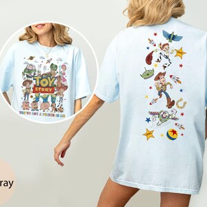 May include: Light blue t-shirt featuring a colorful graphic of Toy Story characters, including Woody, Buzz Lightyear, and others. The front of the shirt displays the Toy Story logo and the phrase "You've Got a Friend in Me."