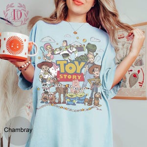 May include: A light blue t-shirt featuring a colorful graphic print of Toy Story characters including Woody, Buzz Lightyear, Jessie, and Rex. The design includes the Toy Story logo and the text You've Got A Friend In Me surrounded by stars.