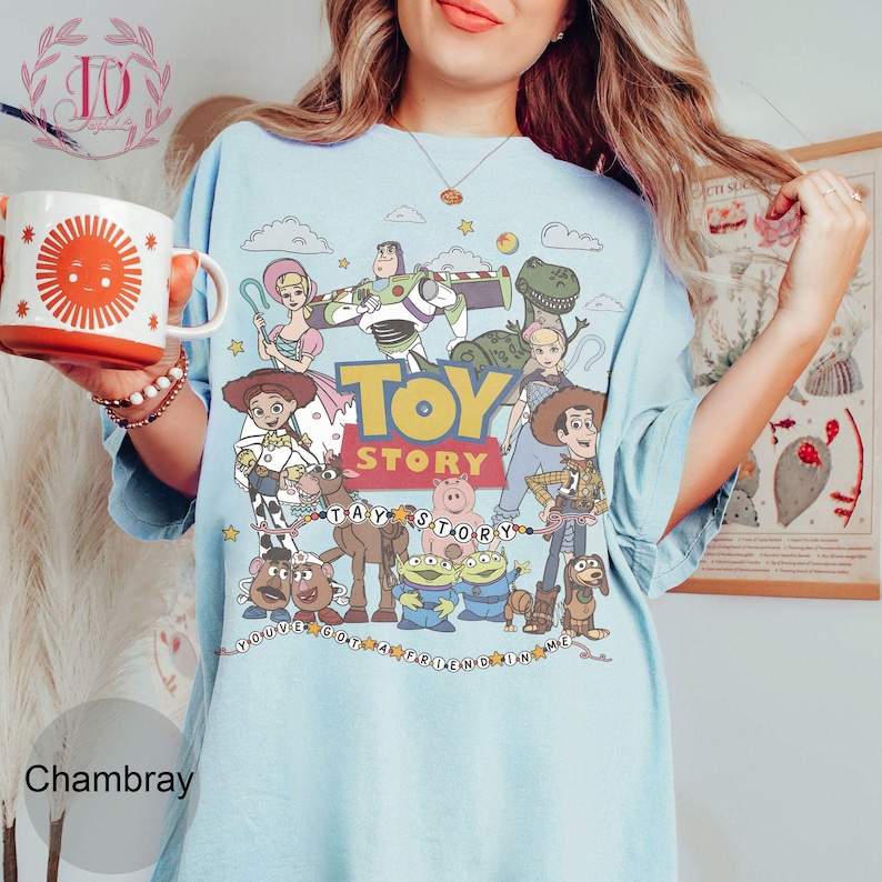May include: A light blue t-shirt featuring a colorful graphic print of Toy Story characters including Woody, Buzz Lightyear, Jessie, and Rex. The design includes the Toy Story logo and the text You've Got A Friend In Me surrounded by stars.