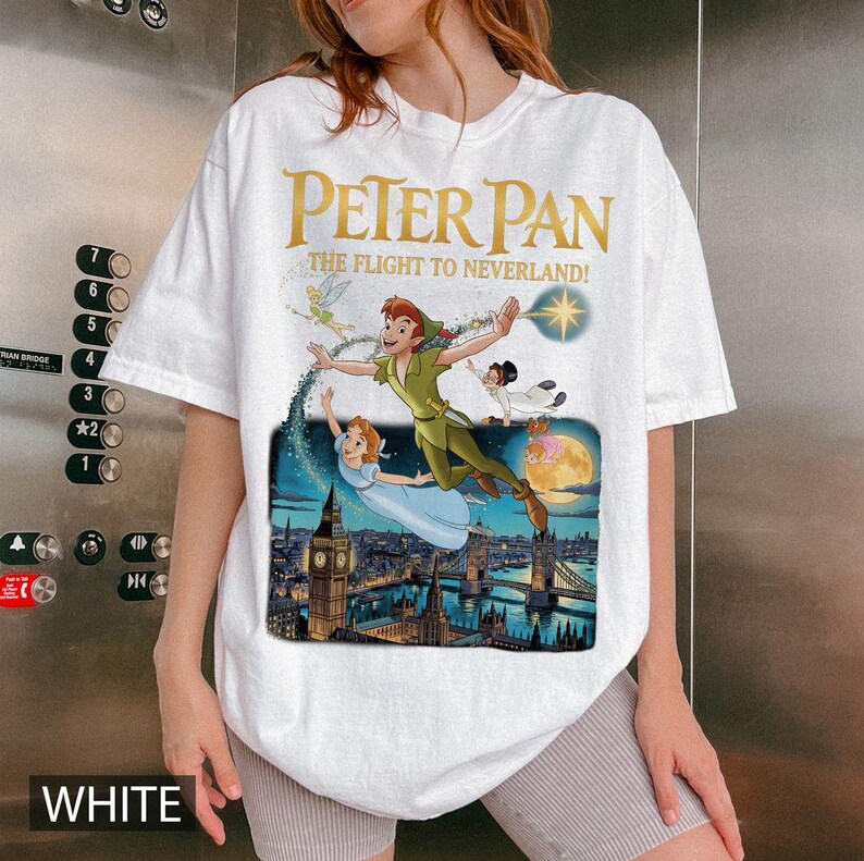 May include: White t-shirt with a vibrant graphic of Peter Pan, Wendy, and other characters soaring above London. The text "PETER PAN THE FLIGHT TO NEVERLAND!" is in gold. The word "WHITE" is printed at the bottom of the shirt.