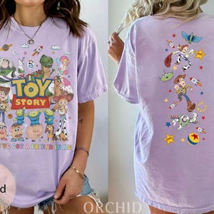 May include: A light purple t-shirt featuring characters from the animated film "Toy Story." The front of the shirt displays the movie title and various characters. The back of the shirt has additional character graphics. The text "YOU'VE GOT A FRIEND IN ME" is also on the front.