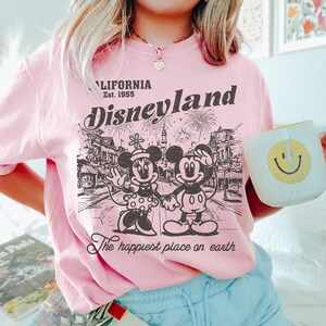 May include: Pink Disneyland graphic t-shirt featuring Mickey and Minnie Mouse, fireworks, and the text "California Est. 1955" and "The happiest place on earth". A white mug with a yellow smiley face is held in the image.