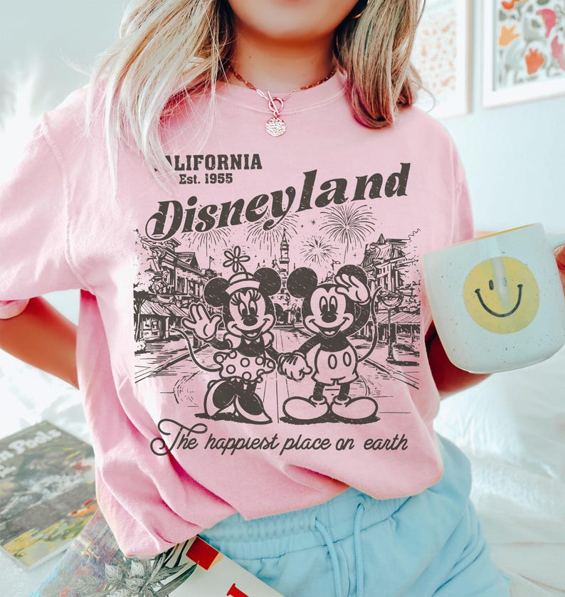 May include: Pink Disneyland graphic t-shirt featuring Mickey and Minnie Mouse, fireworks, and the text "California Est. 1955" and "The happiest place on earth". A white mug with a yellow smiley face is held in the image.