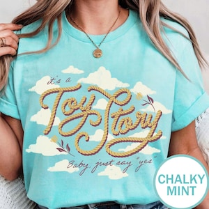 May include: A chalky mint t-shirt featuring a graphic of white clouds and the text "it's a Toy Story" written in a yellow rope-style font, with the additional text "Baby, just say "yes"" printed below in a dark brown cursive script.