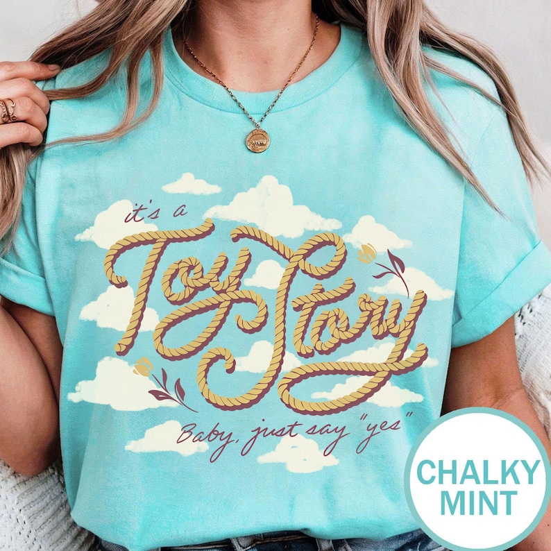 May include: A chalky mint t-shirt featuring a graphic of white clouds and the text "it's a Toy Story" written in a yellow rope-style font, with the additional text "Baby, just say "yes"" printed below in a dark brown cursive script.