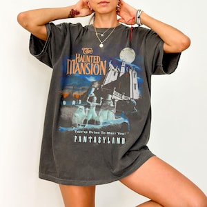 May include: Dark gray t-shirt featuring a graphic from "The Haunted Mansion" ride at Disneyland. The design includes the attraction's name, a spooky mansion, and ghostly figures. The text "They're Dying To Meet You! FANTASYLAND" is also included.