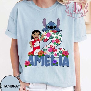 Op de afbeelding: Lichtblauw T-shirt met een kleurrijke afbeelding van Stitch en een meisje, met het nummer 2 en de naam "AMELIA" in blauw. Het nummer 2 is versierd met een bloemenpatroon en bloemen.
