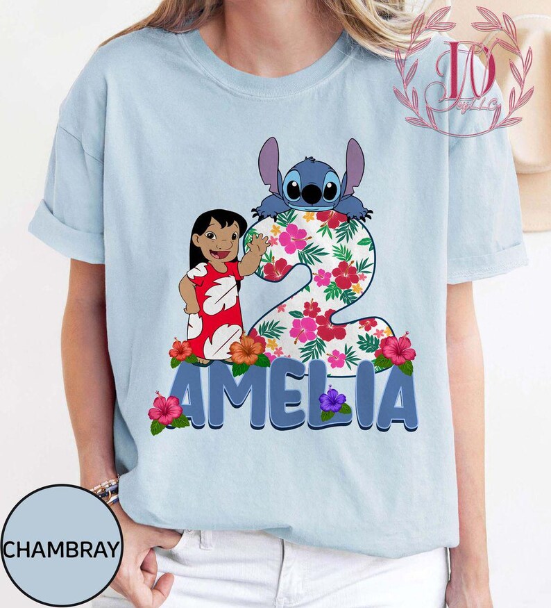 Op de afbeelding: Lichtblauw T-shirt met een kleurrijke afbeelding van Stitch en een meisje, met het nummer 2 en de naam "AMELIA" in blauw. Het nummer 2 is versierd met een bloemenpatroon en bloemen.