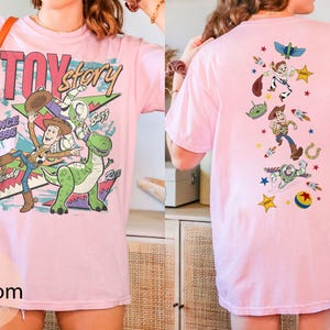 May include: A pale pink t-shirt with a vibrant Toy Story design. The front shows Woody, Buzz Lightyear, and Rex, with the text "Toy Story Since 1995". The back of the shirt features a vertical arrangement of the characters and related imagery.