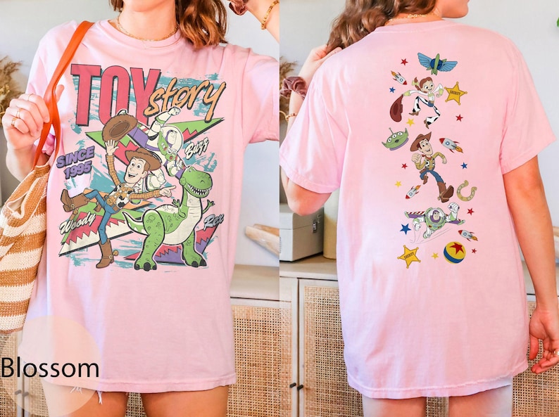 May include: A pale pink t-shirt with a vibrant Toy Story design. The front shows Woody, Buzz Lightyear, and Rex, with the text "Toy Story Since 1995". The back of the shirt features a vertical arrangement of the characters and related imagery.