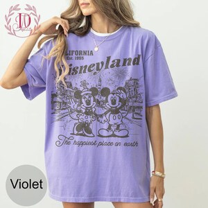 May include: Violet t-shirt with a graphic of Mickey and Minnie Mouse in front of a Disneyland scene. The shirt includes the text "California Est. 1955 Disneyland" and "The happiest place on earth."