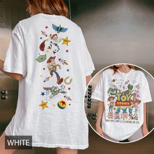 May include: White t-shirt featuring colorful graphics of characters from the animated film "Toy Story." The back of the shirt displays Woody, Buzz Lightyear, and other characters with stars and rockets. The front of the shirt has the words "Toy Story" and "You've Got a Friend in Me."
