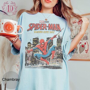 Puede incluir: Camiseta azul claro con un gr&aacute;fico de Spider-Man de estilo vintage. El dise&ntilde;o incluye el texto "MARVEL STUDIOS SPIDER-MAN BRAND NEW DAY" y una ilustraci&oacute;n de Spider-Man contra un horizonte urbano. Una taza blanca y naranja con un dise&ntilde;o de sol se sostiene en la mano izquierda.