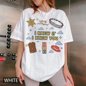 Puede incluir: Una camiseta blanca de manga corta con un estampado gr&aacute;fico que incluye una estrella de sheriff amarilla, una pulsera de cuentas, botas de vaquero marrones, un sombrero rojo y bloques de letras. El texto dice "infinity and beyond", "YOU'VE GOT A FRIEND IN ME" e "I KNEW IT I KNEW YOU".