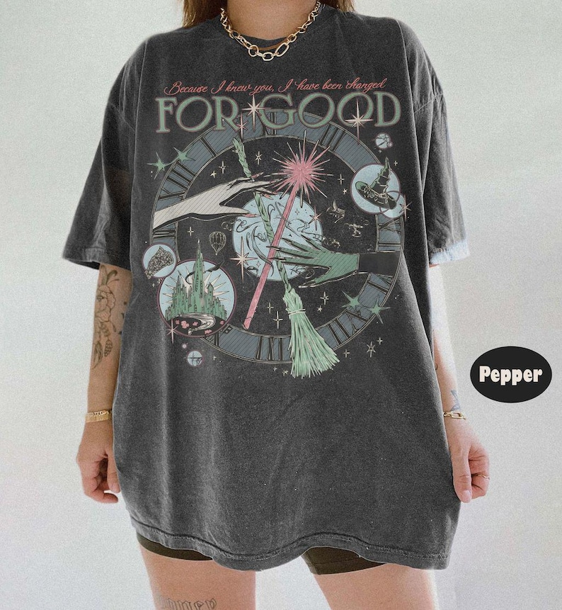 Puede incluir: Camiseta gris oscuro con un dise&ntilde;o gr&aacute;fico que presenta una esfera de reloj, manos, una varita m&aacute;gica y el texto "FOR GOOD" y "Because I knew you, I have been changed." El dise&ntilde;o incluye un castillo y elementos caprichosos.