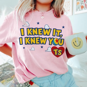 May include: A light pink t-shirt featuring the yellow and blue text I KNEW IT, I KNEW YOU, with a colourful ball icon replacing the letter O, and a red heart containing the yellow letters TS, surrounded by small blue stars and white clouds.