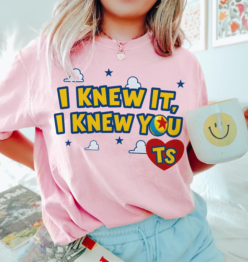May include: A light pink t-shirt featuring the yellow and blue text I KNEW IT, I KNEW YOU, with a colourful ball icon replacing the letter O, and a red heart containing the yellow letters TS, surrounded by small blue stars and white clouds.