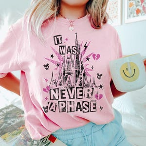 May include: A light pink t-shirt with a graphic design featuring a castle, lightning bolts, stars, and the text "IT WAS NEVER A PHASE". A white mug with a yellow smiley face is held in the image.