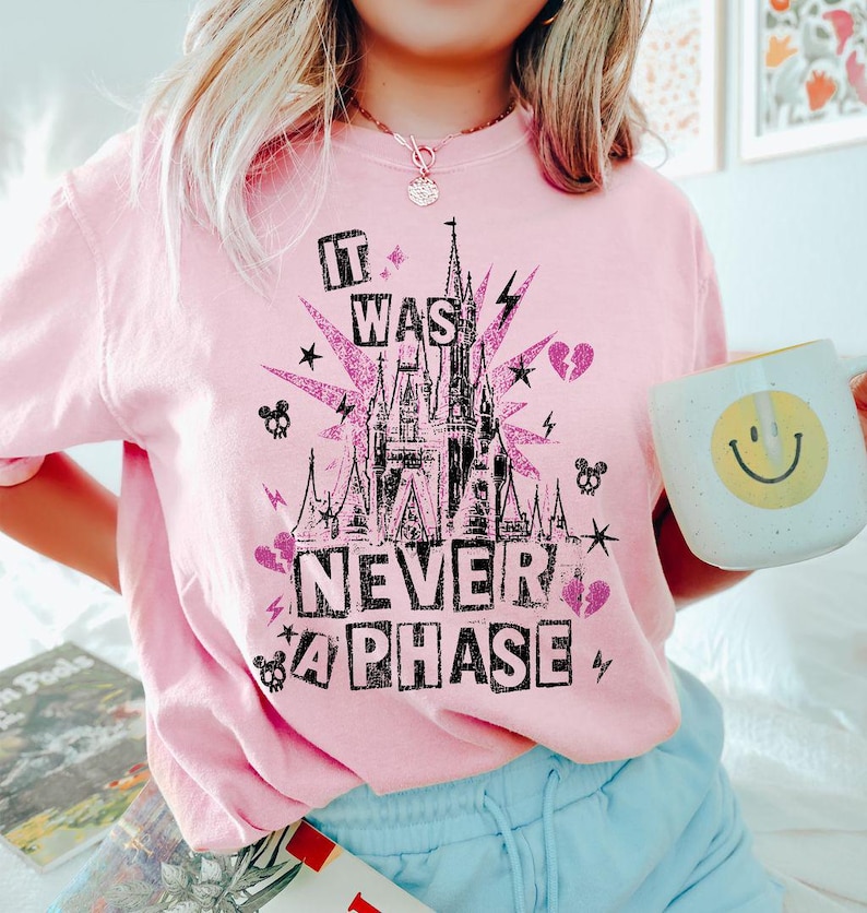 May include: A light pink t-shirt with a graphic design featuring a castle, lightning bolts, stars, and the text "IT WAS NEVER A PHASE". A white mug with a yellow smiley face is held in the image.