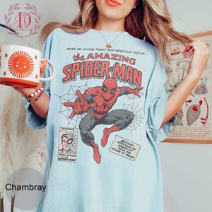 Puede incluir: Camiseta azul claro con un gr&aacute;fico de c&oacute;mic vintage de Spider-Man en rojo y negro, con el texto "The Amazing Spider-Man". La camiseta tambi&eacute;n tiene paneles y texto de c&oacute;mic adicionales. En la imagen se sostiene una taza blanca y naranja con un dise&ntilde;o de sol.
