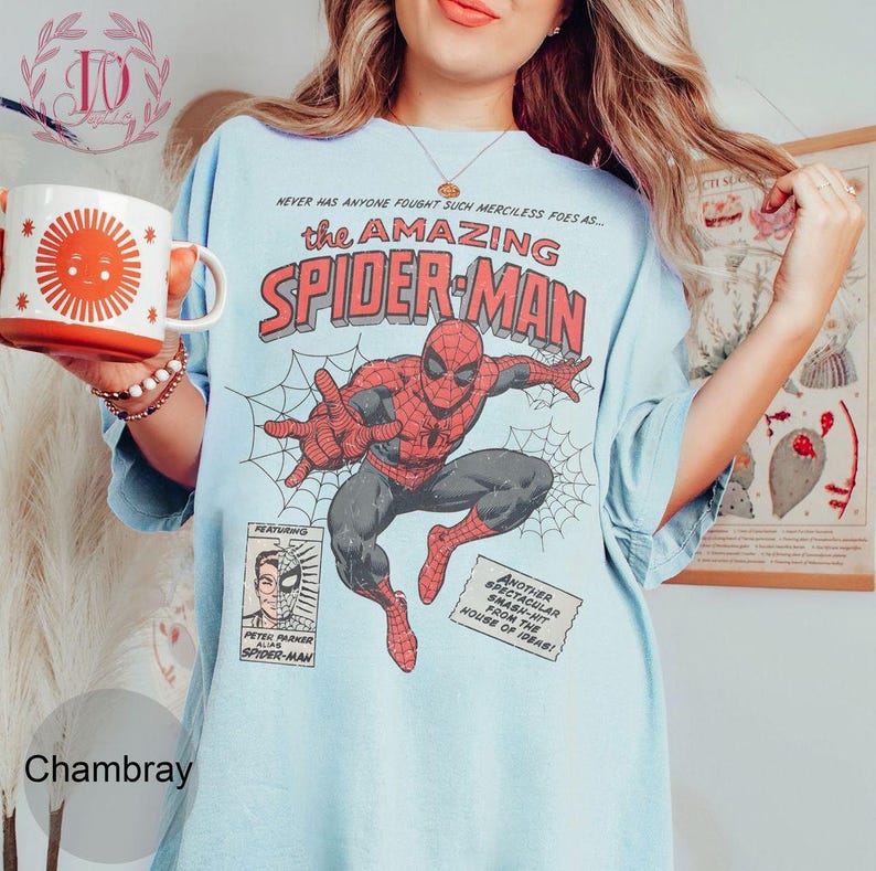 Puede incluir: Camiseta azul claro con un gr&aacute;fico de c&oacute;mic vintage de Spider-Man en rojo y negro, con el texto "The Amazing Spider-Man". La camiseta tambi&eacute;n tiene paneles y texto de c&oacute;mic adicionales. En la imagen se sostiene una taza blanca y naranja con un dise&ntilde;o de sol.