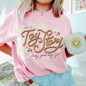 May include: A light pink short-sleeve t-shirt featuring a graphic design with the text "it's a Toy Story" written in a brown rope-style font, accompanied by the phrase "Baby, just say "yes"" and small floral illustrations over pale yellow clouds.