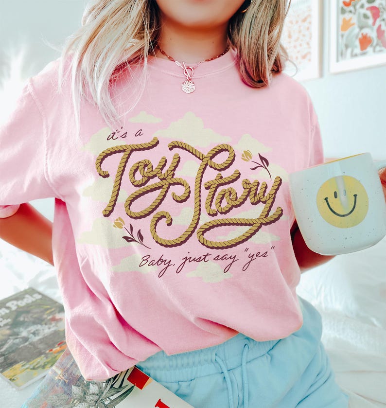 May include: A light pink short-sleeve t-shirt featuring a graphic design with the text "it's a Toy Story" written in a brown rope-style font, accompanied by the phrase "Baby, just say "yes"" and small floral illustrations over pale yellow clouds.
