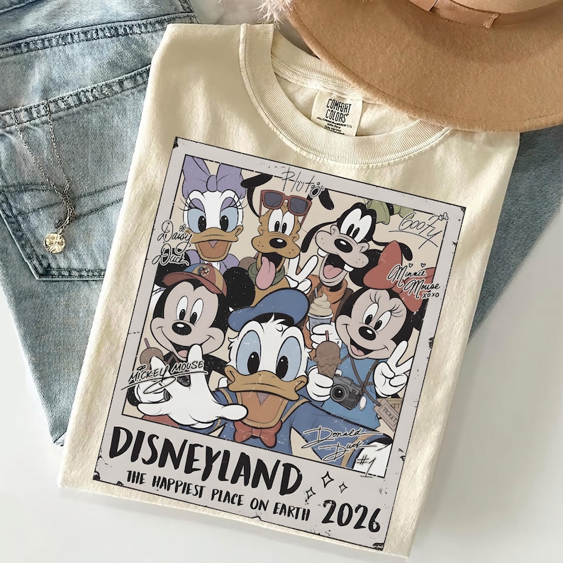 K&ouml;nnte beinhalten: Cremefarbenes T-Shirt mit einem Vintage-Disneyland-Grafikdesign mit Micky Maus, Minnie Maus, Donald Duck und anderen Disney-Figuren. Die Grafik enth&auml;lt den Text "Disneyland" und "The Happiest Place on Earth 2026."